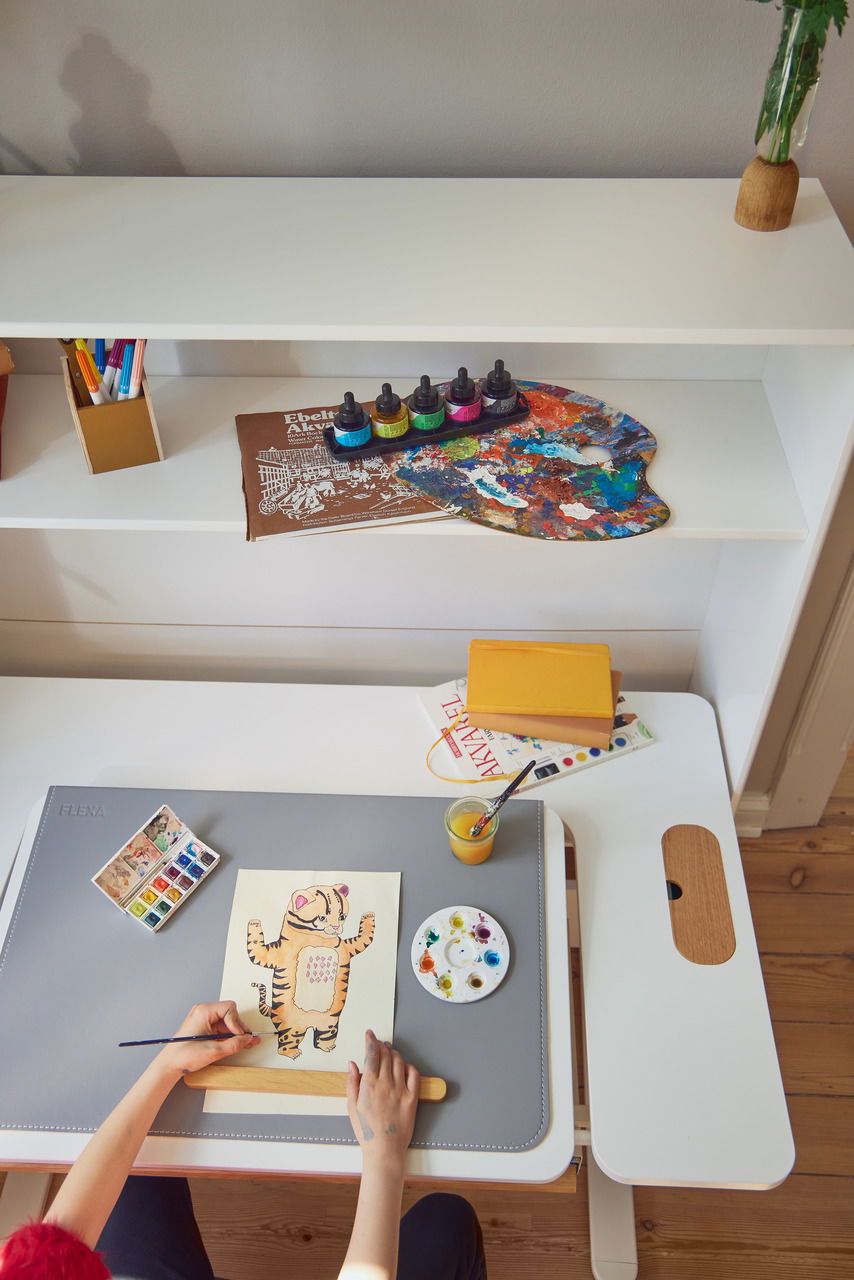 Vogelperspektive auf einen Schreibtisch mit grauer Schreibtisch-Auflage, auf der ein Kind malt. Daneben sind Wasserfarben, ein Pinsel und ein Glas mit Wasser zu sehen.
