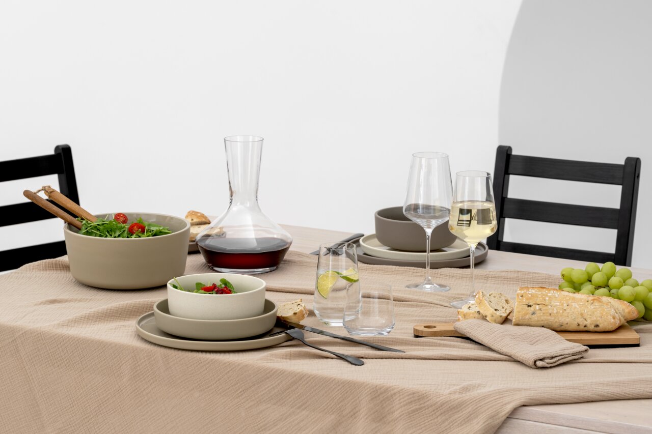 Een stijlvol gedekte tafel met in het midden een wijnkaraf gevuld met rode wijn. De karaf staat op een beige tafelkleed, omringd door serviesgoed, glazen en een selectie gerechten. De foto toont de tafel in zijaanzicht.