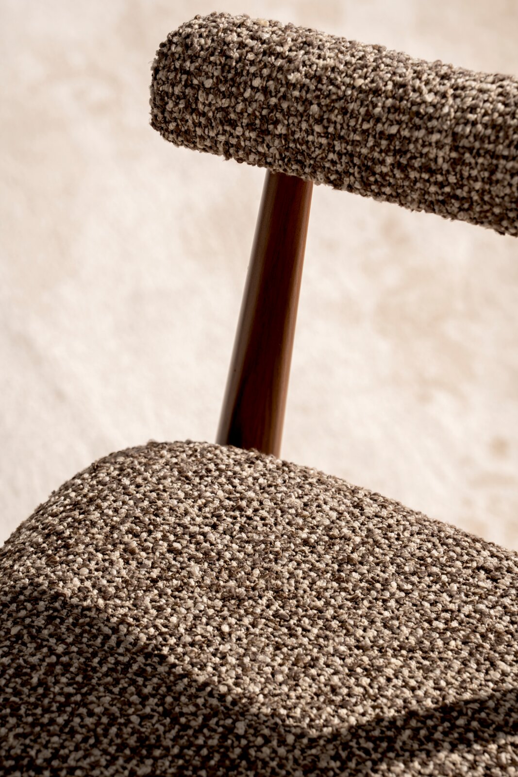 Nahaufnahme eines gepolsterten Stuhls mit grob gewebtem, meliertem Stoffbezug in Beige-Braun und dunkler Holzstrebe; Perspektive: Detailansicht schräg von der Seite.