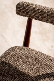 Nahaufnahme eines gepolsterten Stuhls mit grob gewebtem, meliertem Stoffbezug in Beige-Braun und dunkler Holzstrebe; Perspektive: Detailansicht schräg von der Seite.