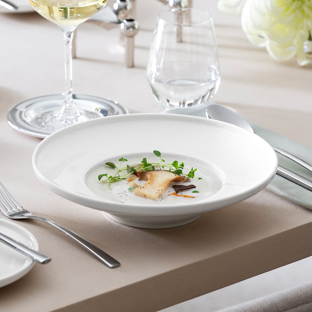 Villeroy & Boch Soepbord AFINA Eleganter weißer Suppenteller Afina 25cm, gefüllt mit einer cremigen Suppe und dekoriert mit frischen Kräutern, auf einem gedeckten Tisch von der Seite fotografiert.