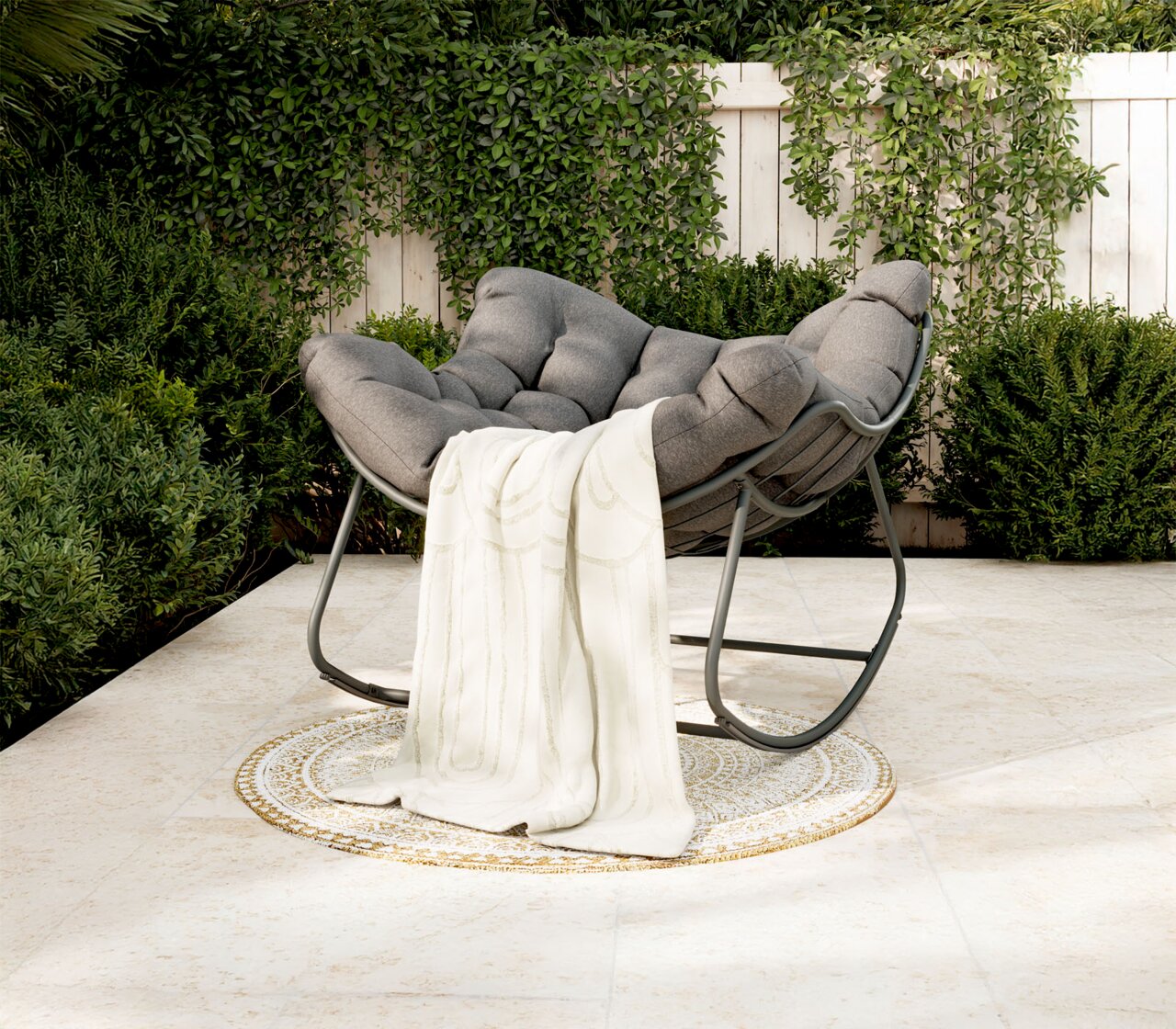 Ambia Garden Schaukelsessel FARO Gemütlicher grauer Schaukelsessel mit weicher Polsterung und weißer Decke, auf einer Terrasse mit grüner Pflanzenkulisse, seitliche Perspektive.