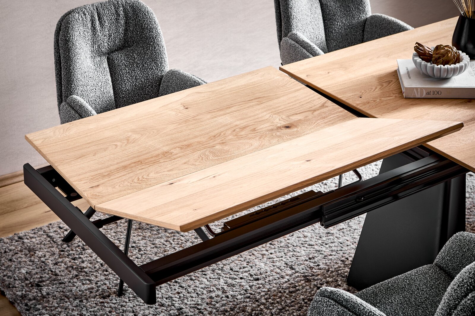 Ausziehbarer Esstisch mit heller Holzplatte und schwarzem Metallgestell, teilweise ausgezogen mit sichtbarer Einlegeplatte, Ansicht schräg von oben (3/4-Perspektive) im Essbereich mit grauen Polsterstühlen.