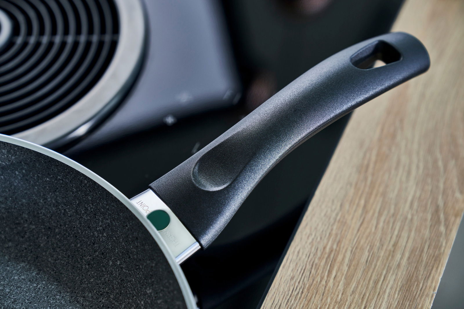 Nahaufnahme eines Pfannengriffs aus schwarzem Kunststoff, der an einer Pfanne befestigt ist, mit einem Kochfeld im Hintergrund.