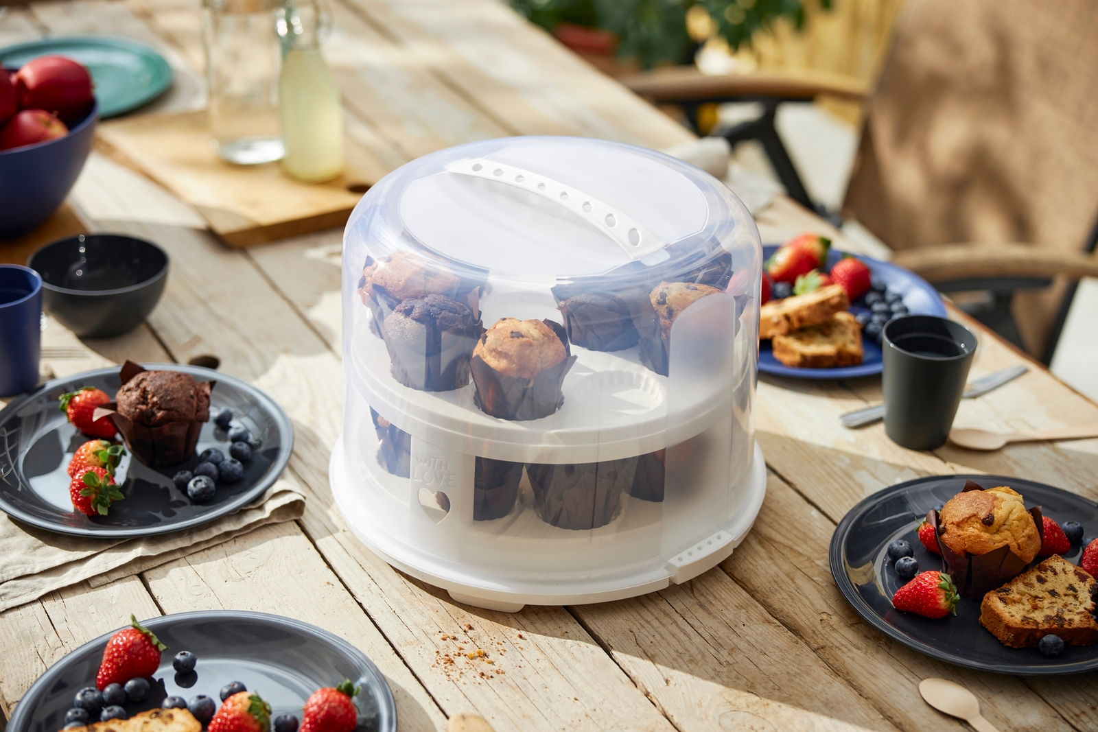 rotho Tortenglocke XL FRESH Tortenglocke XL 26 cm mit Tray, gefüllt mit Muffins, auf einem Holztisch im Freien, umgeben von Tellern mit Beeren und Gebäck, aus seitlicher Perspektive