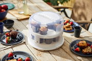 Tortenglocke XL 26 cm mit Tray, gefüllt mit Muffins, auf einem Holztisch im Freien, umgeben von Tellern mit Beeren und Gebäck, aus seitlicher Perspektive