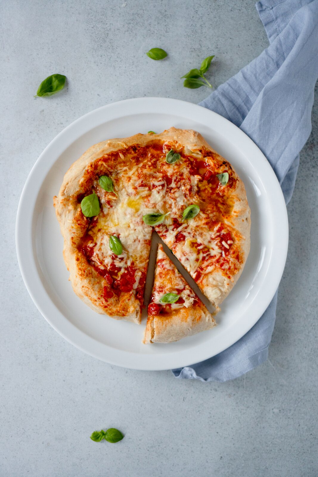 Hausgemachte Margherita-Pizza mit Tomatensauce, geschmolzenem Käse und frischem Basilikum auf weißem Teller, ein Stück herausgeschnitten, Draufsicht (von oben).