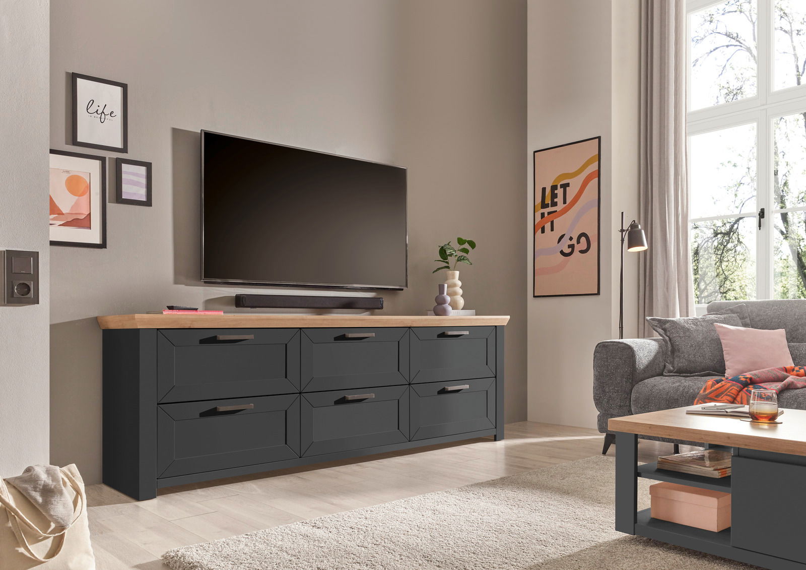 Set one by Musterring Sideboard SET ONE YORK Modernes Sideboard in dunklem Holz mit sechs Schubladen, platziert in einem stilvoll eingerichteten Wohnzimmer. Perspektive von vorne links.