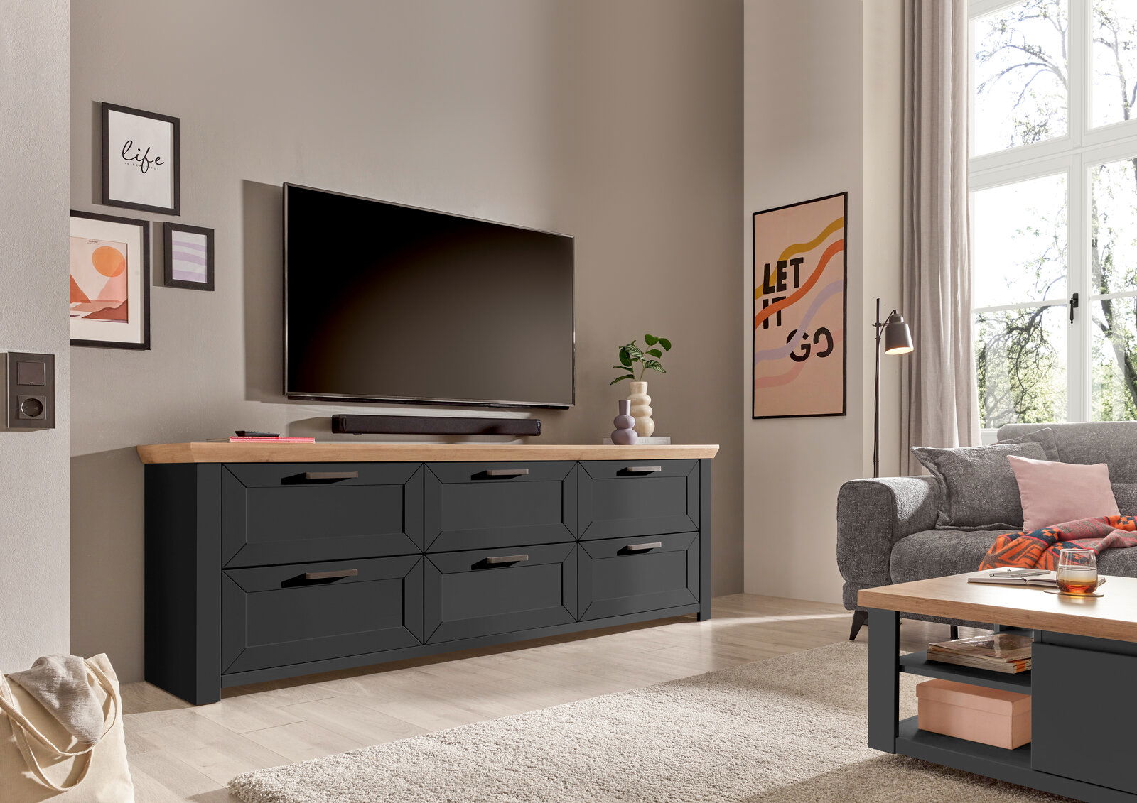 Set one by Musterring Sideboard SET ONE YORK Modernes Sideboard in dunklem Holz mit sechs Schubladen, platziert in einem stilvoll eingerichteten Wohnzimmer. Perspektive von vorne links.