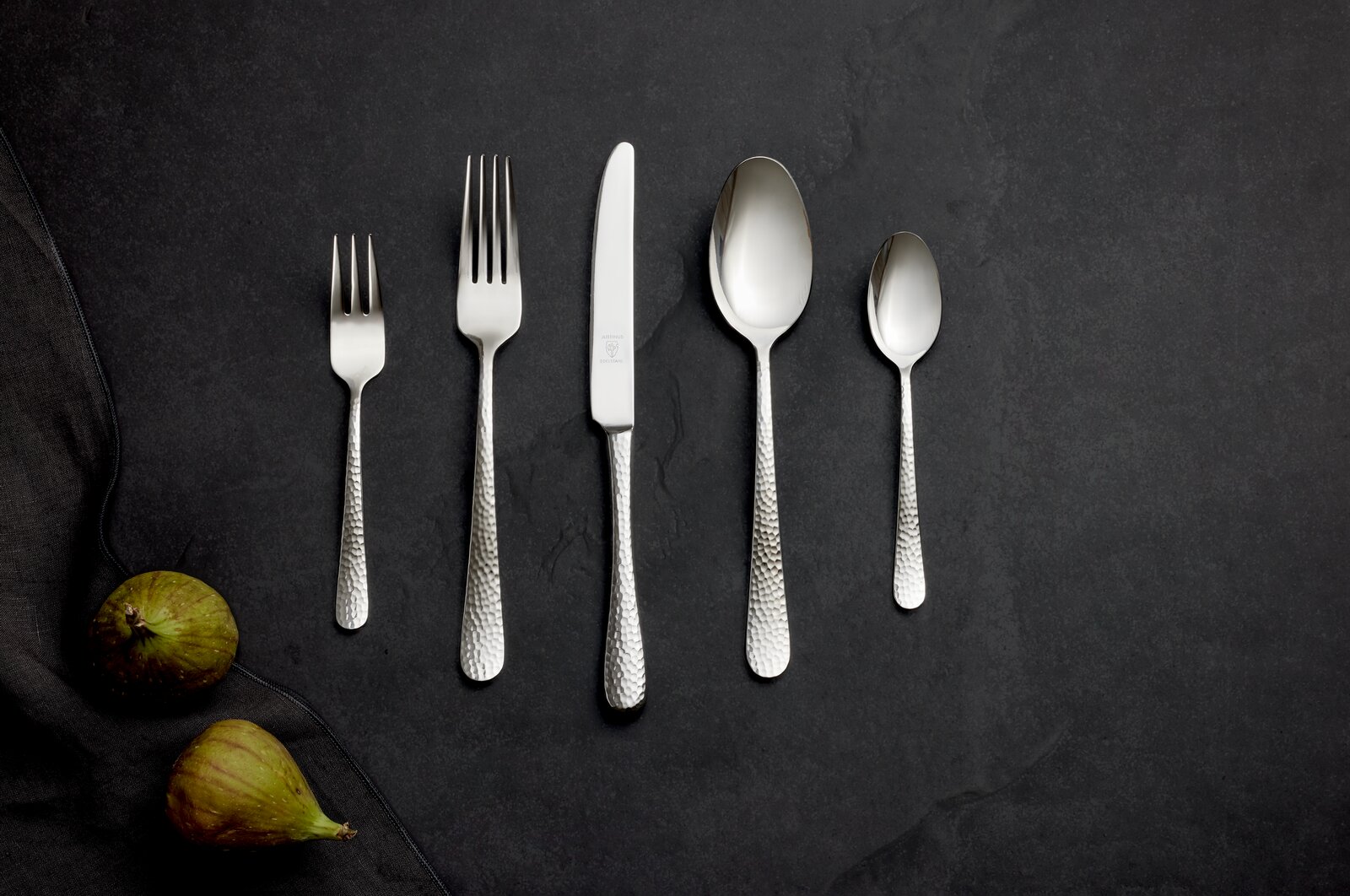 Besteckset aus Edelstahl mit gehämmertem Griff, bestehend aus Gabel, Messer, Löffel und Teelöffel, auf dunklem Hintergrund von oben fotografiert.