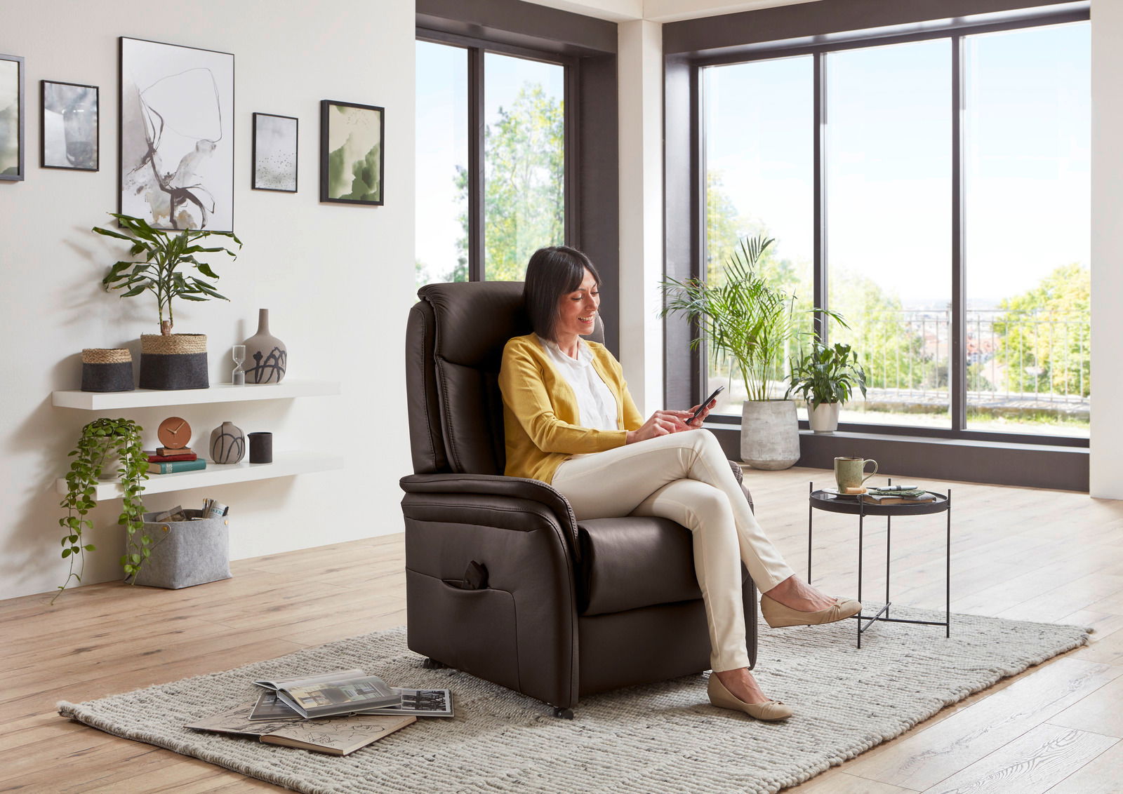 Frau sitzt entspannt in einem braunen Relax-Sessel in einem modernen Wohnzimmer mit großen Fenstern und Pflanzen, seitliche Perspektive.
