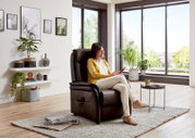 Een vrouw zit ontspannen in een bruine relaxfauteuil in een moderne woonkamer met grote ramen en planten, gezien vanuit een zijdelingse hoek.