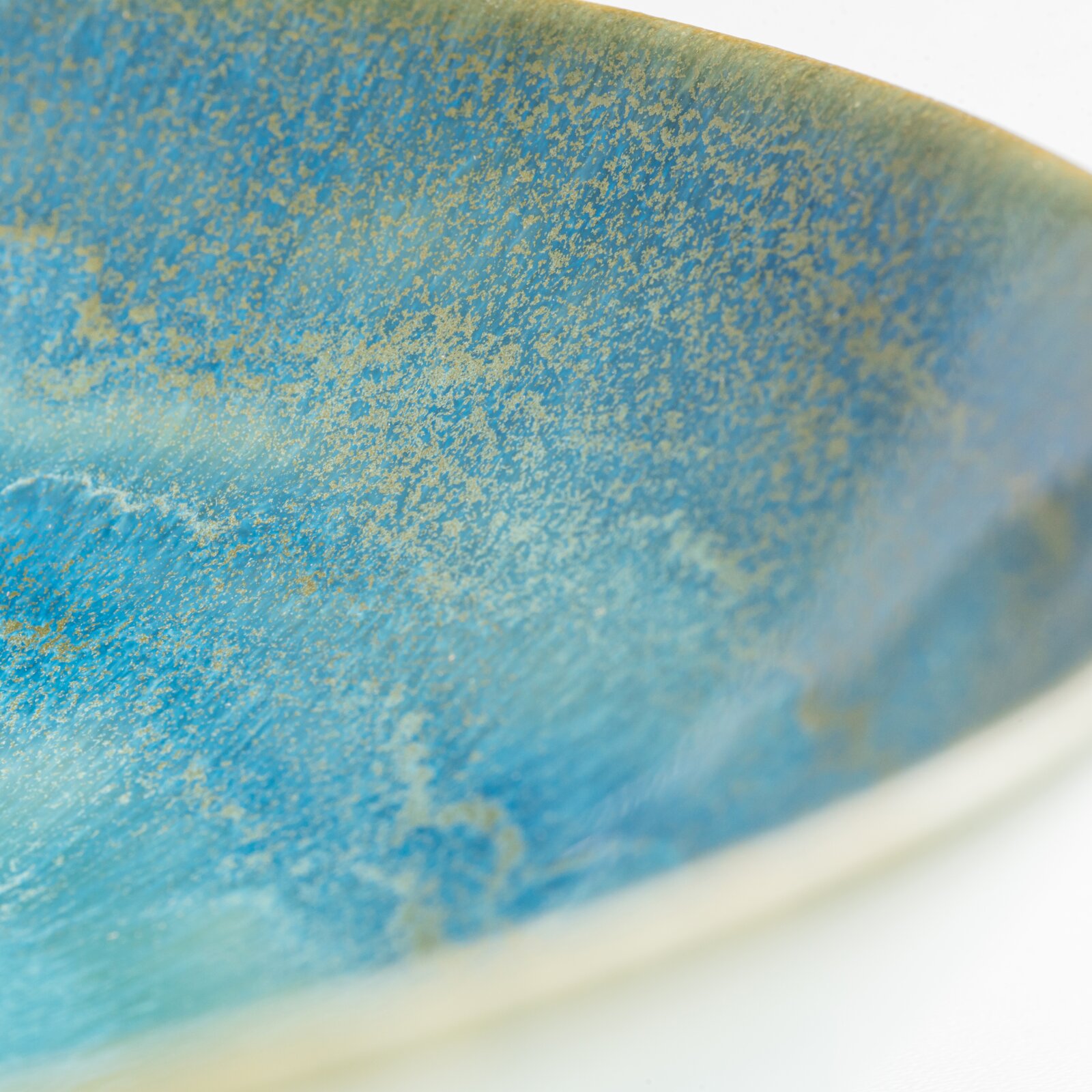 Makro-Nahaufnahme aus schräger Seitenperspektive einer blau-türkis glasierten Keramikschale mit goldenen Sprenkeln und sichtbarem Rand