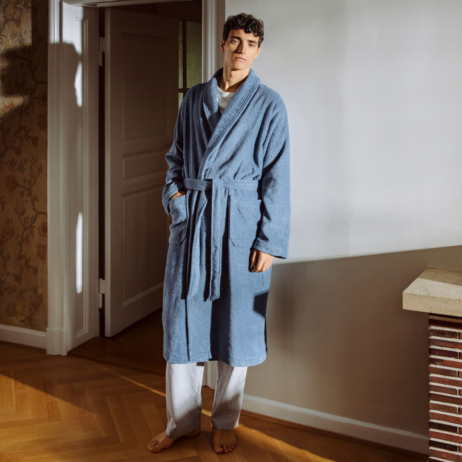 Mann in einem blauen Bademantel mit Schalkragen steht in einem gemütlichen Raum, frontal zur Kamera.