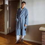 Mann in einem blauen Bademantel mit Schalkragen steht in einem gemütlichen Raum, frontal zur Kamera.