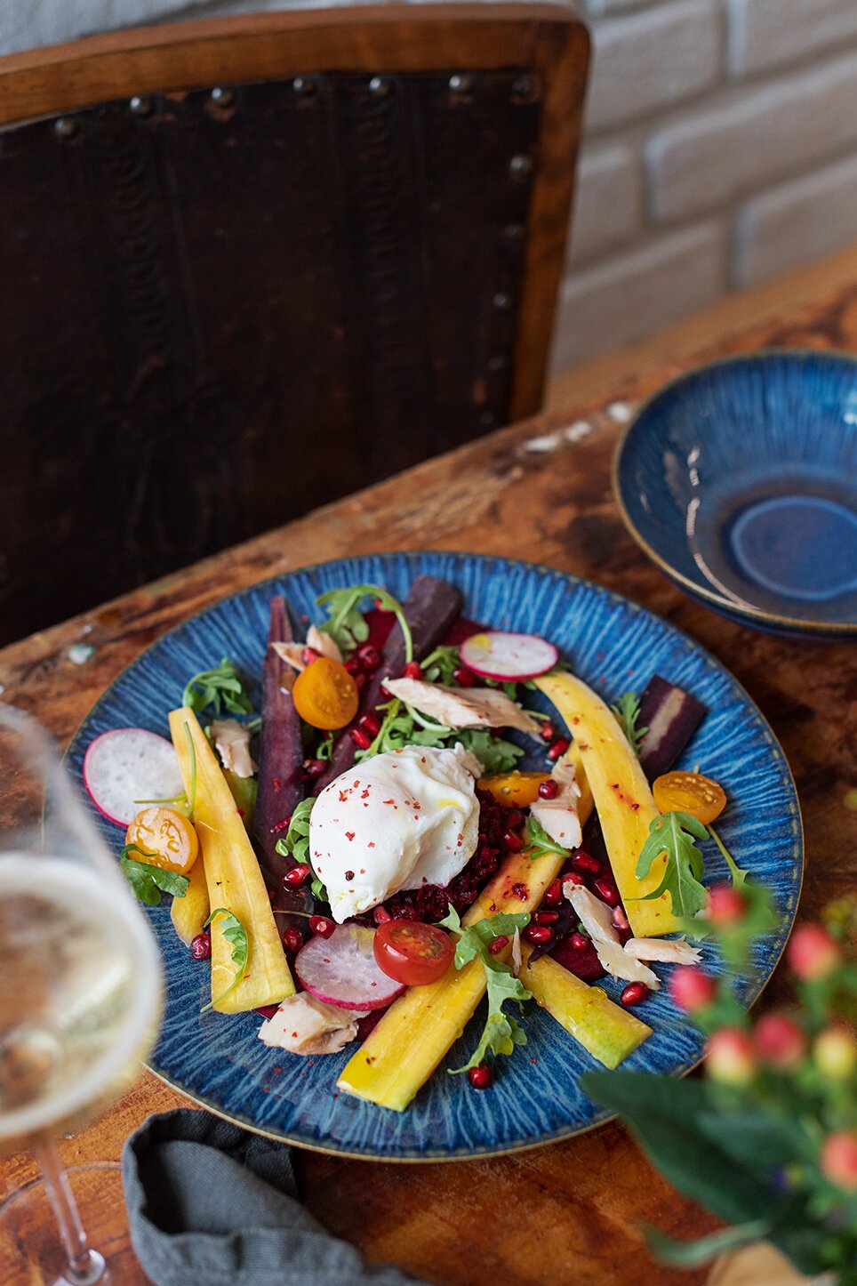 Schräg von oben fotografierter, farbenfroher Salat auf blauem Teller mit Burrata, Rucola, roter Bete, Radieschen, Granatapfelkernen und gelben Gemüsestreifen auf rustikalem Holztisch.