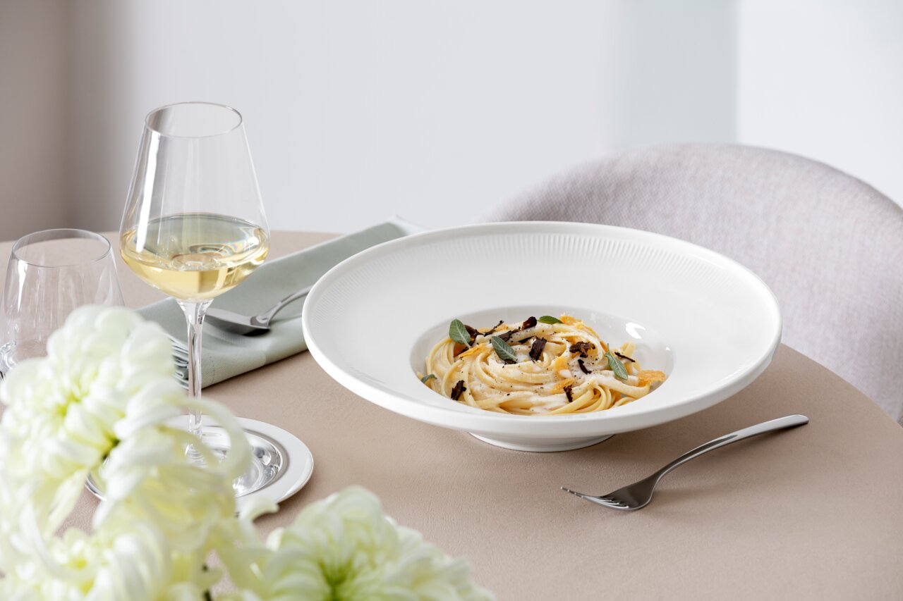 Elegant wit soepbord Afina met een diameter van 29,2 cm, gevuld met pasta, op een gedekte tafel met een wijnglas en bloemen, zijaanzicht.