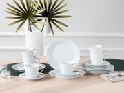 Weißes Porzellan-Geschirrset mit Tassen, Untertassen und Tellern auf hellem Holztisch, frontal in leichter Draufsicht fotografiert, im minimalistischen Interieur mit Vase und Palmblättern im Hintergrund
