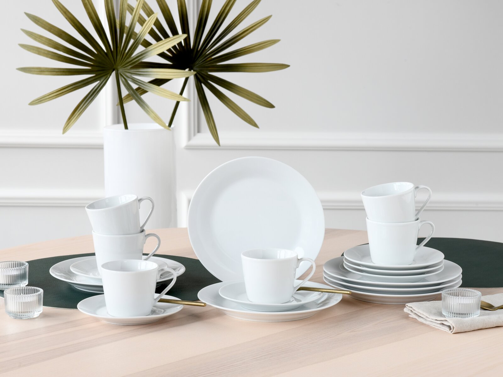 Weißes Porzellan-Geschirrset mit Tassen, Untertassen und Tellern auf hellem Holztisch, frontal in leichter Draufsicht fotografiert, im minimalistischen Interieur mit Vase und Palmblättern im Hintergrund