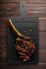 Schneidebrett mit Rillen, darauf ein saftiges, gegrilltes Tomahawk-Steak mit Gewürzen, aus der Vogelperspektive fotografiert.