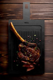 Schneidebrett mit Rillen, darauf ein saftiges, gegrilltes Tomahawk-Steak mit Gewürzen, aus der Vogelperspektive fotografiert.