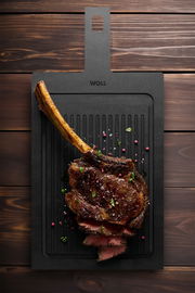 Schneidebrett mit Rillen, darauf ein saftiges, gegrilltes Tomahawk-Steak mit Gewürzen, aus der Vogelperspektive fotografiert.