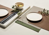 Tischset in verschiedenen Farben, darunter Braun, Grau und Grün, mit einem Teller und einer Schale, dekoriert mit Olivenzweigen, aus der Vogelperspektive fotografiert.