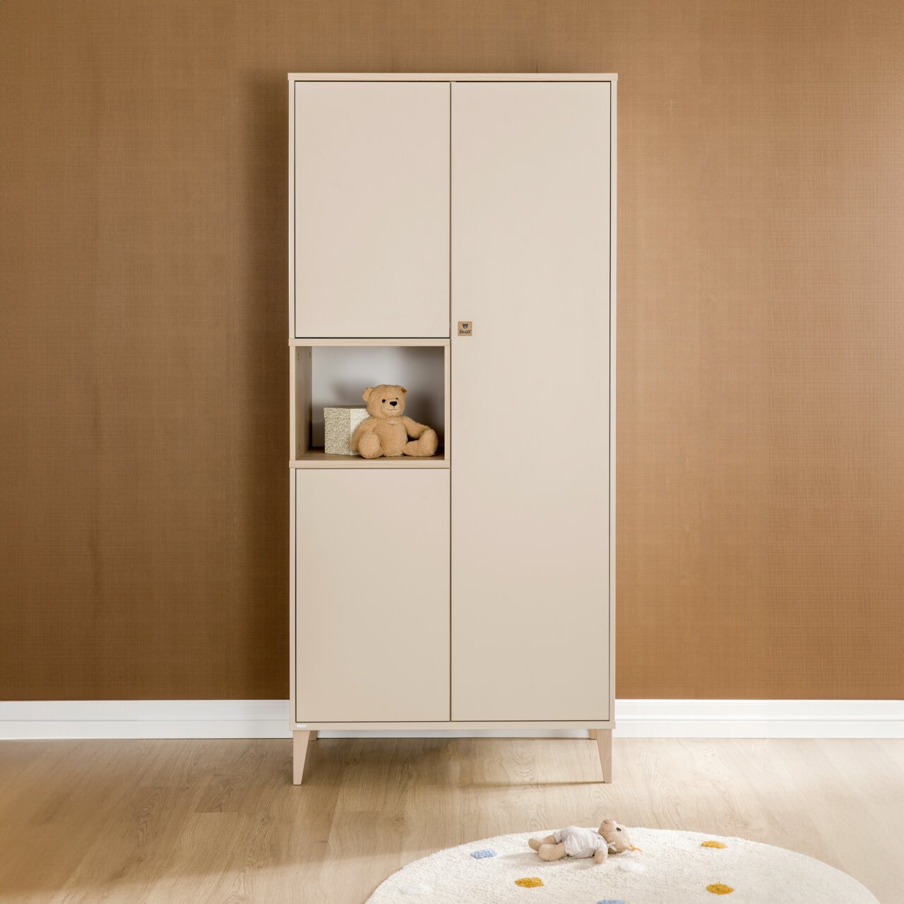 Frontalansicht eines dreitürigen Kleiderschranks der Marke Paidi, Modell MILA&BEN, in einem warmen Beige-Ton. Der Schrank hat ein offenes Fach mit einem Teddybär und einer kleinen Box.
