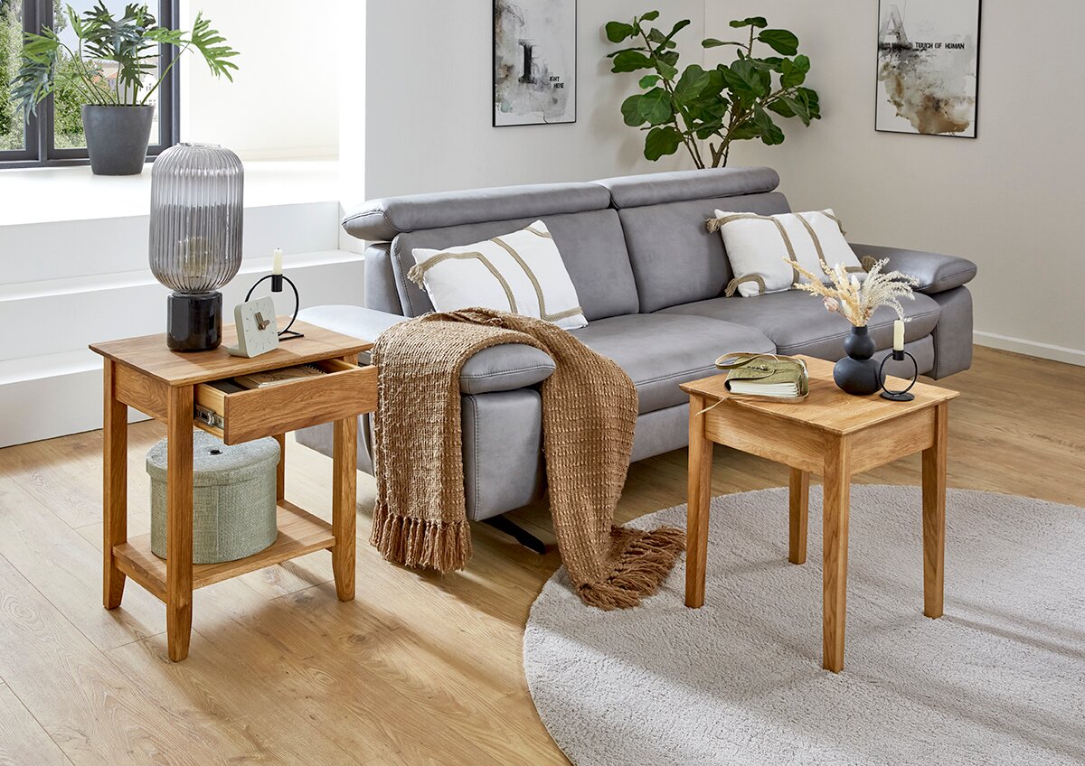 Zwei kleine Beistelltische aus hellem Massivholz im Wohnzimmer neben einem grauen Sofa, links mit Schublade und Ablagefach, fotografiert in leichter Schrägansicht von vorne.