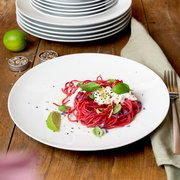 Ein weißer Teller aus einem 18-teiligen Geschirrset, präsentiert mit einer Portion roter Pasta, garniert mit Basilikum und Limette, auf einem Holztisch. Perspektive von oben.