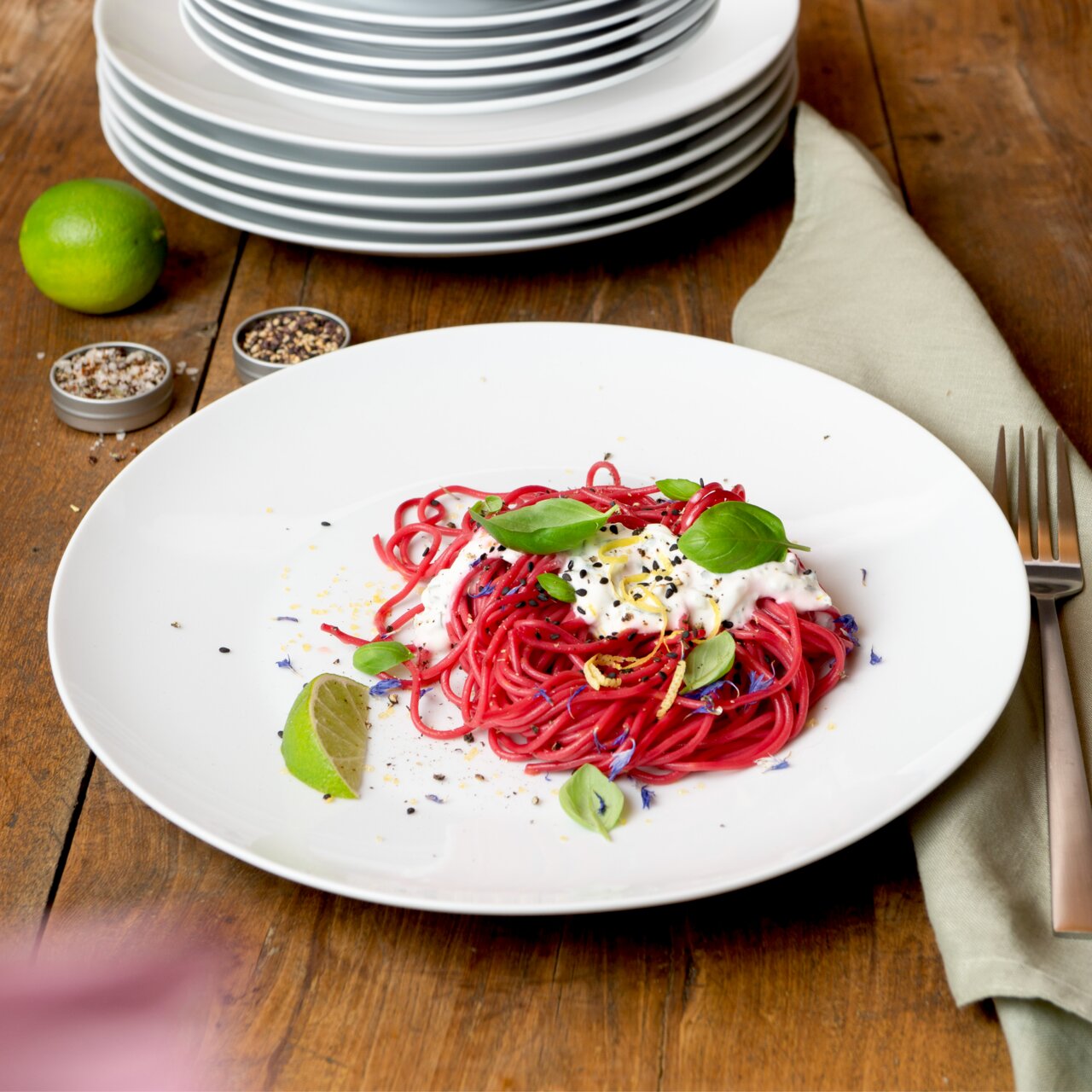 Ein weißer Teller aus einem 18-teiligen Geschirrset, präsentiert mit einer Portion roter Pasta, garniert mit Basilikum und Limette, auf einem Holztisch. Perspektive von oben.
