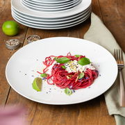 Ein weißer Teller aus einem 18-teiligen Geschirrset, präsentiert mit einer Portion roter Pasta, garniert mit Basilikum und Limette, auf einem Holztisch. Perspektive von oben.