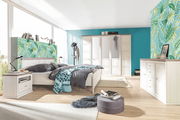 Helles Schlafzimmer mit einem 5-türigen Drehtürenschrank, einem großen Bett, einer Nachtkommode und einem Sideboard in Holzoptik. Die Perspektive zeigt den Raum von der Seite mit Blick auf das Bett und den Schrank.