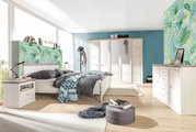 Helles Schlafzimmer mit einem 5-türigen Drehtürenschrank, einem großen Bett, einer Nachtkommode und einem Sideboard in Holzoptik. Die Perspektive zeigt den Raum von der Seite mit Blick auf das Bett und den Schrank.