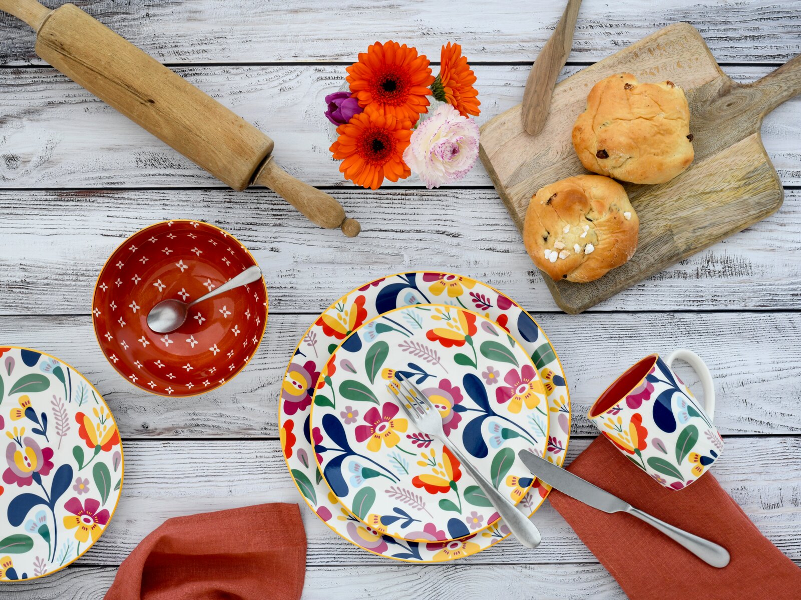 Draufsicht auf buntes, florales Geschirr-Set auf weißer Holzoberfläche: Teller mit Besteck, passende Tasse, rote Schale mit Löffel, Holzbrett mit Brötchen, Nudelholz und orangefarbene Blumen.