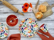 Draufsicht auf buntes, florales Geschirr-Set auf weißer Holzoberfläche: Teller mit Besteck, passende Tasse, rote Schale mit Löffel, Holzbrett mit Brötchen, Nudelholz und orangefarbene Blumen.