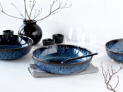 Blaue, gesprenkelte Keramikschale mit schwarzem Löffel auf grauem Leinentuch, umgeben von passendem Geschirr, Gläsern und schwarzer Vase auf weißem Tisch, fotografiert aus leicht erhöhter Frontperspektive.