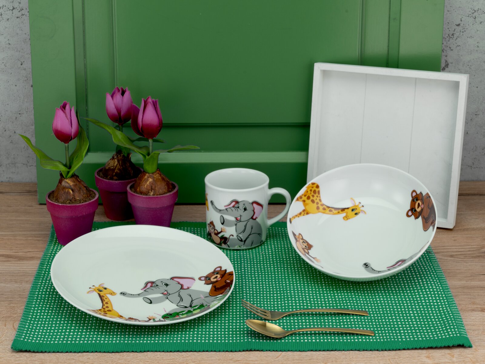 Kinder-Geschirrset aus weißem Porzellan mit Tiermotiven (Teller, Schüssel und Tasse) auf grünem Platzset, frontal aus leicht erhöhter Perspektive fotografiert; im Hintergrund pinke Tulpen im Topf und ein weißes Tablett.
