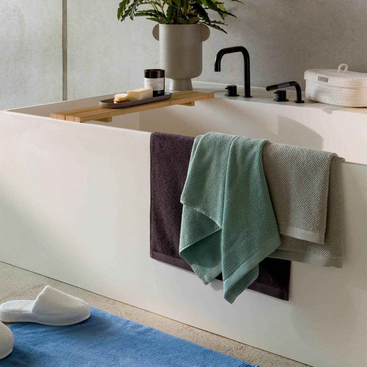 Blick auf ein modernes Badezimmer mit einer Badewanne, über der ein Holzbrett mit Seife und einer Kerze liegt. Drei Handtücher in verschiedenen Farben hängen über dem Wannenrand. Im Vordergrund sind Hausschuhe auf einem blauen Teppich zu sehen.
