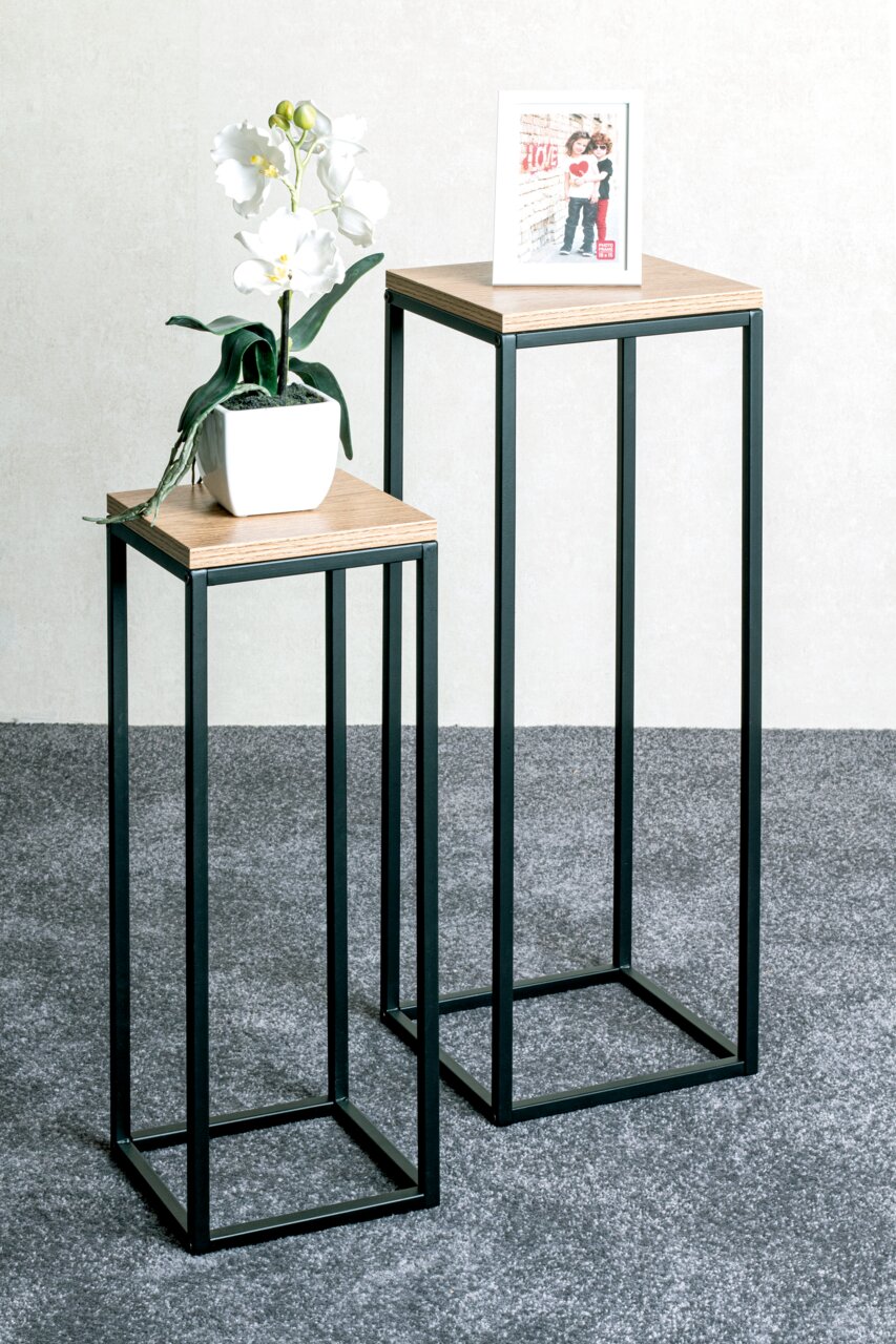 Zwei moderne Beistelltische mit schwarzem Metallrahmen und Holzoberfläche, einer mit einer weißen Orchidee und der andere mit einem Bilderrahmen, aus seitlicher Perspektive fotografiert.