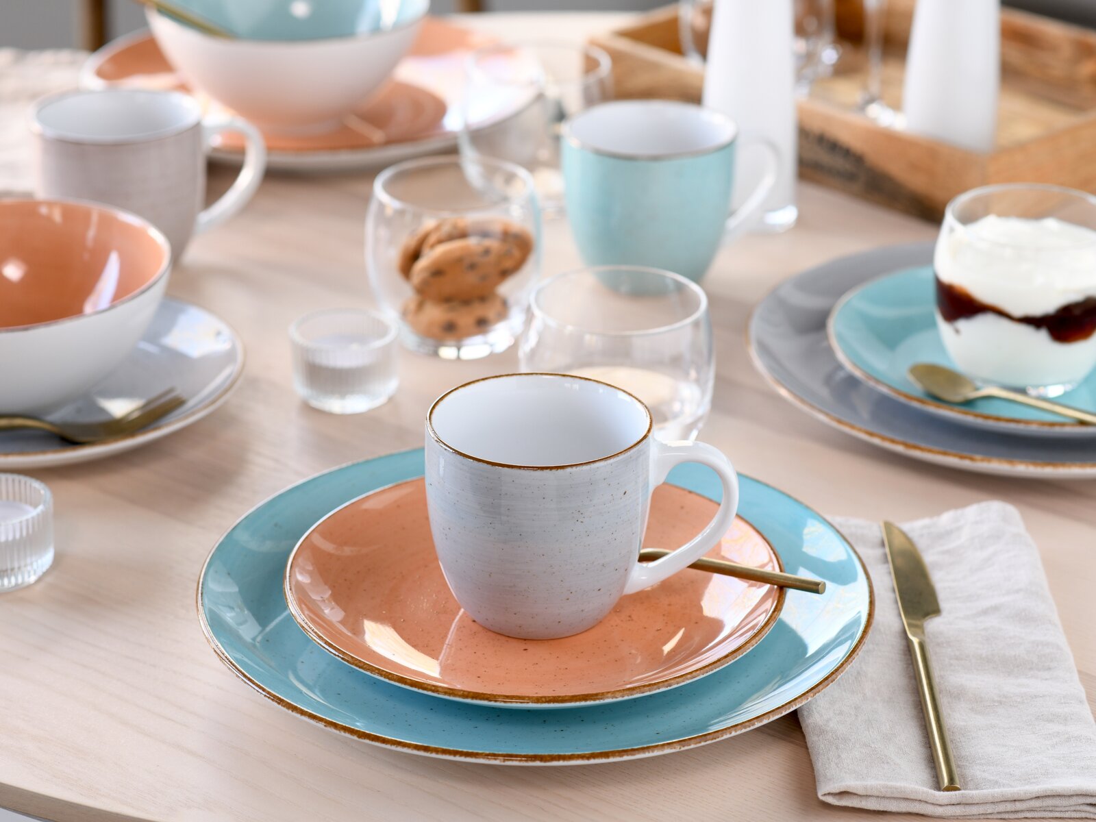 Gedeckter Frühstückstisch mit Keramikgeschirr in Pastellfarben: weiße Tasse auf lachsfarbener Untertasse und türkisfarbenem Teller, goldfarbenes Besteck, Gläser und Schälchen im Hintergrund, Perspektive: leicht von oben in schräger Frontansicht.