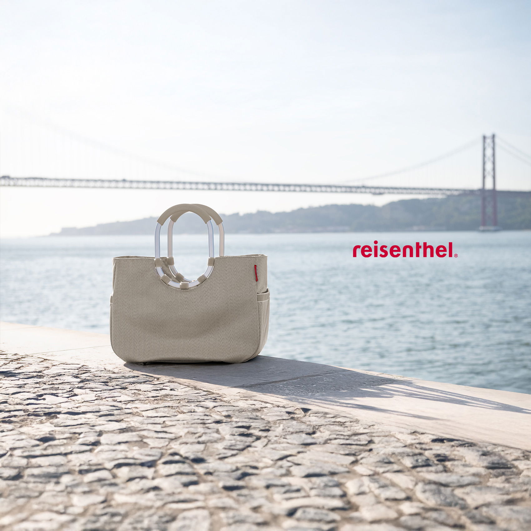 Beige Reisenthel-Tasche am Ufer vor Wasser und Brücke, Logo rechts im Bild