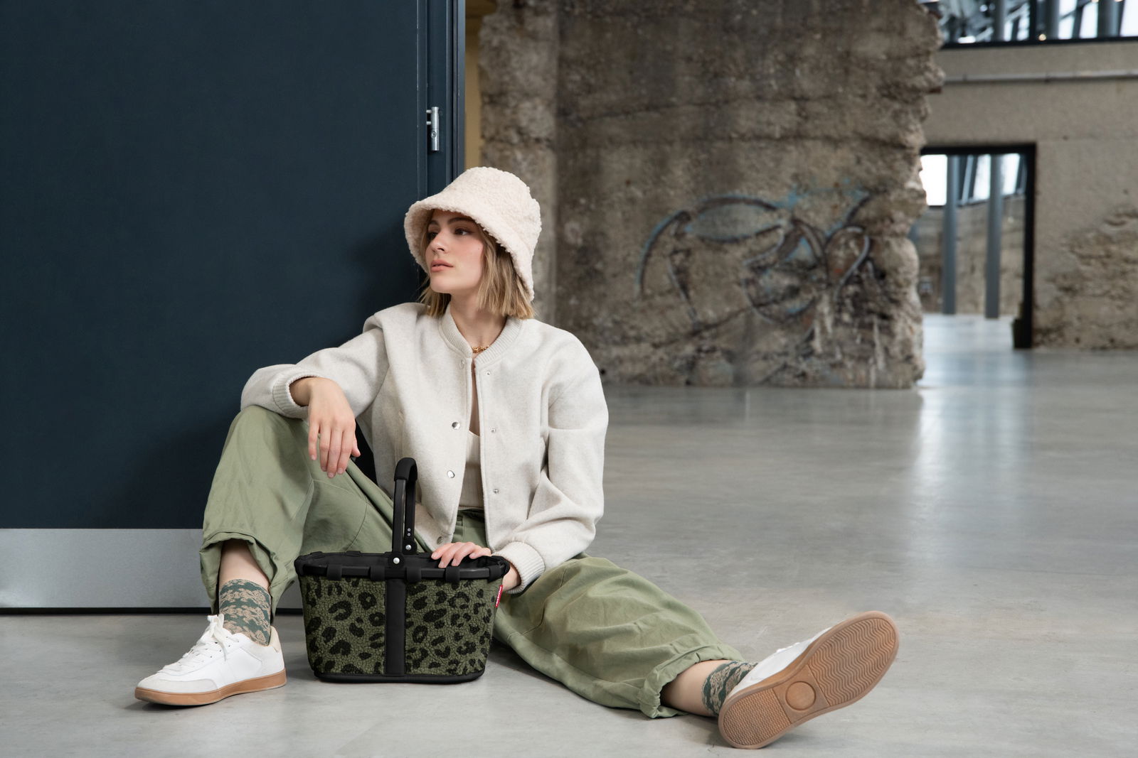 Frau sitzt lässig auf dem Boden in einem modernen, industriellen Raum und hält einen kleinen, grünen Korb mit Leopardenmuster in der Hand. Perspektive von vorne.