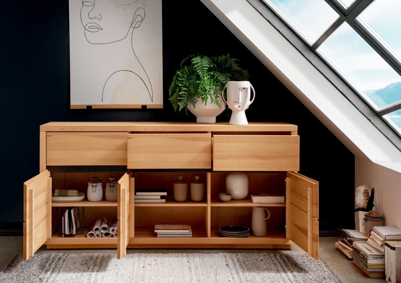 Holz-Sideboard mit offenen Schubladen und Türen, dekoriert mit Pflanzen und Keramik, in einem modernen Raum unter einem Dachfenster, Frontalansicht.