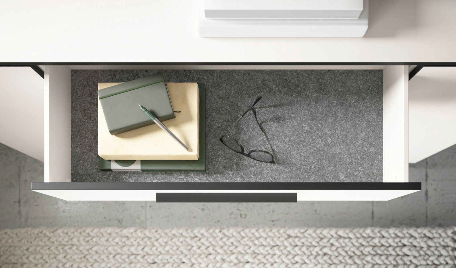 Offene Schublade einer Kommode von oben, gefüllt mit Büchern, einem Stift und einer Brille auf grauem Filz.