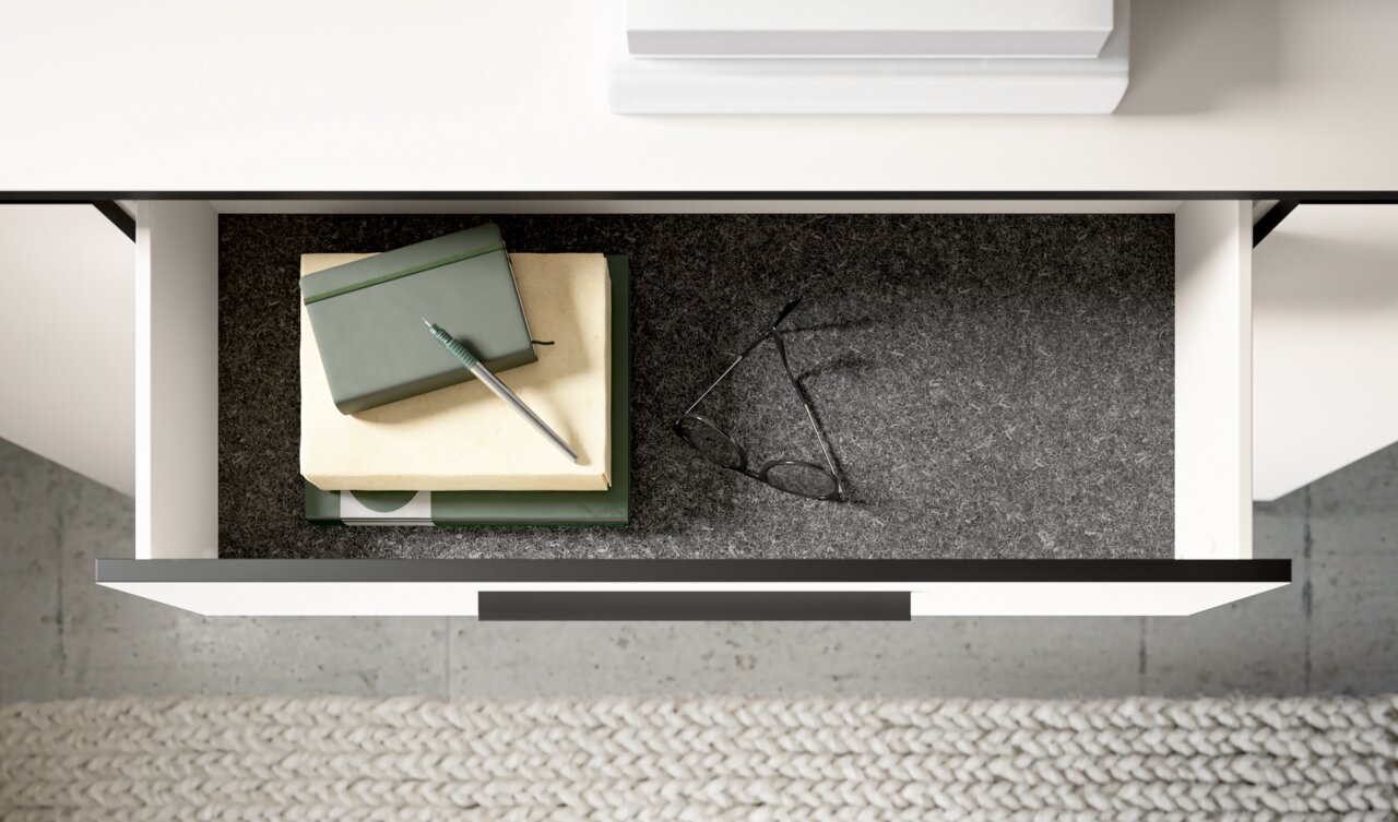 Offene Schublade einer Kommode von oben, gefüllt mit Büchern, einem Stift und einer Brille auf grauem Filz.