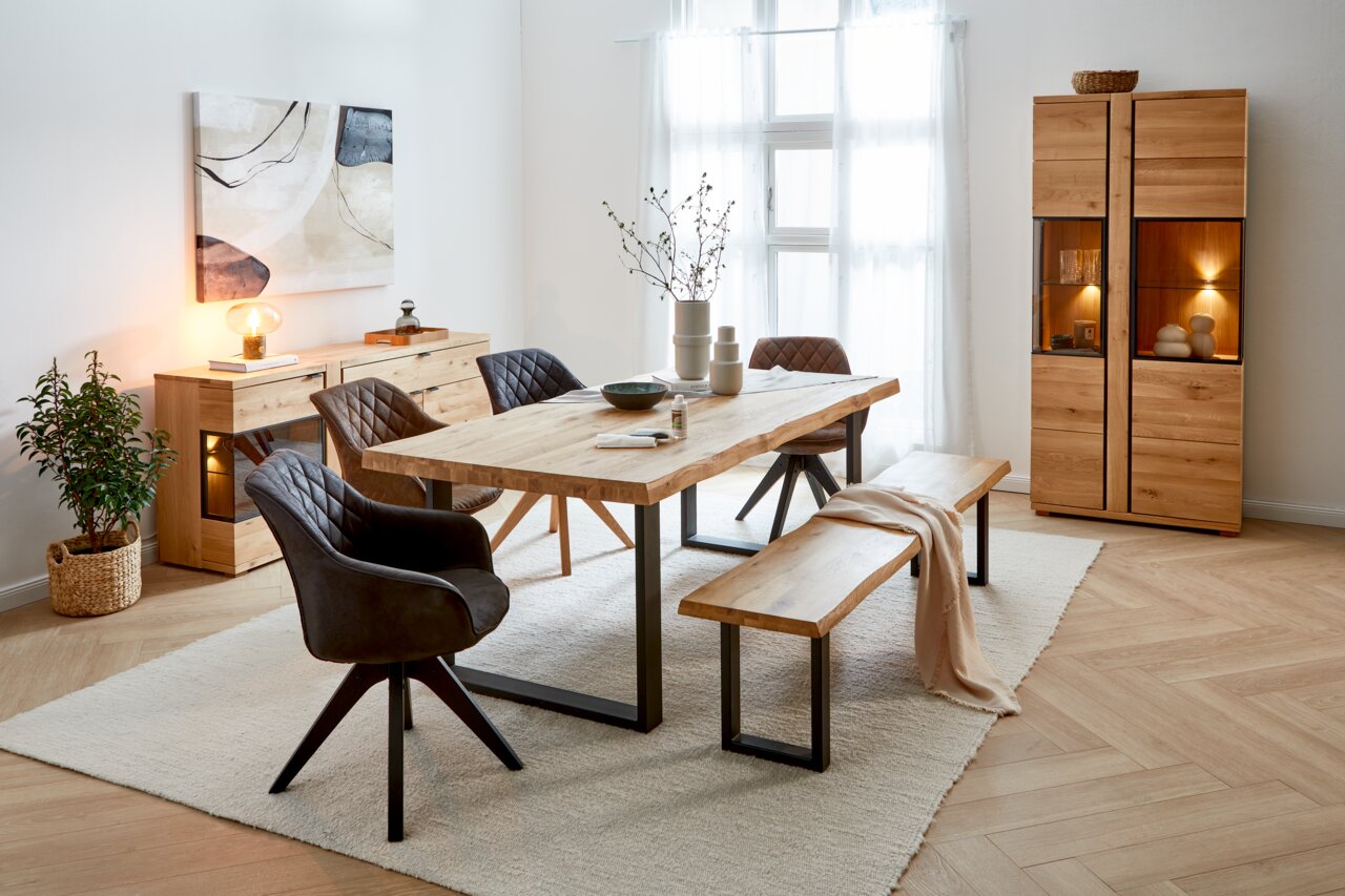 Esszimmer mit modernem Holztisch, umgeben von gepolsterten Stühlen in dunklem Leder und einer Holzbank. Im Hintergrund sind ein Sideboard und ein Schrank aus Holz zu sehen. Die Perspektive zeigt den Raum von der Seite, mit Blick auf den Tisch und die Stüh
