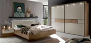 Modernes Schlafzimmer-Set mit Holzbett und gepolstertem Kopfteil, Nachttisch und großem Schwebetürenschrank in Weiß mit Holz-Akzentstreifen und LED-Spots, gezeigt in frontaler leicht seitlicher Perspektive.