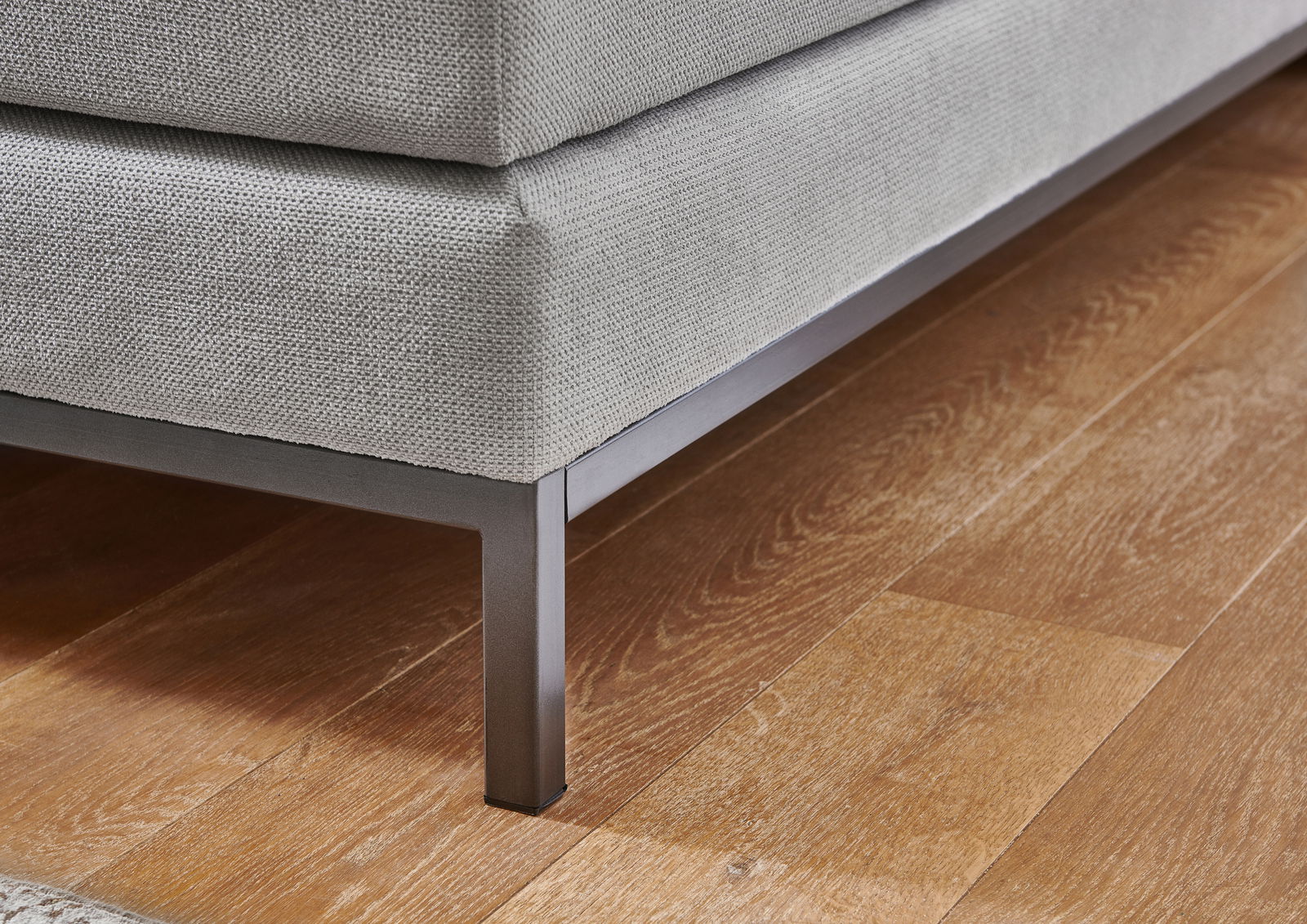 Nahaufnahme eines Boxspringbettes mit grauem Stoffbezug und Metallbeinen auf Holzboden, seitliche Perspektive