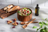 Salatschüssel-Set aus Akazienholz mit frischem Salat, umgeben von einem Holzlöffel-Set, einem Hackblock mit Baguette, Zwiebeln und einer Flasche Olivenöl auf einem hellen Holztisch. Perspektive von oben.